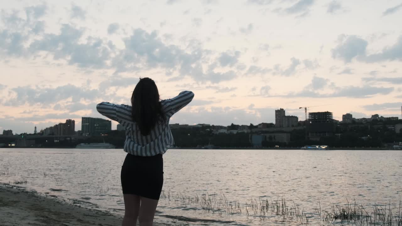 美丽的女孩穿着黑色的裙子和条纹衬衫站在日落的背景下的城市在河边，抿着她的手。后视图。慢动作视频素材