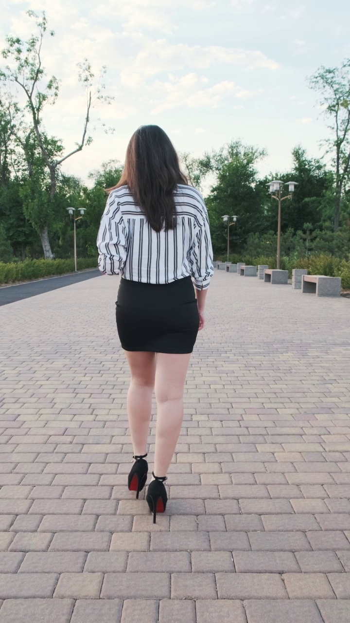 美丽的女孩穿着黑色的高跟鞋，黑色的裙子和条纹衬衫走在公园里。后视图。慢动作，垂直视频视频素材