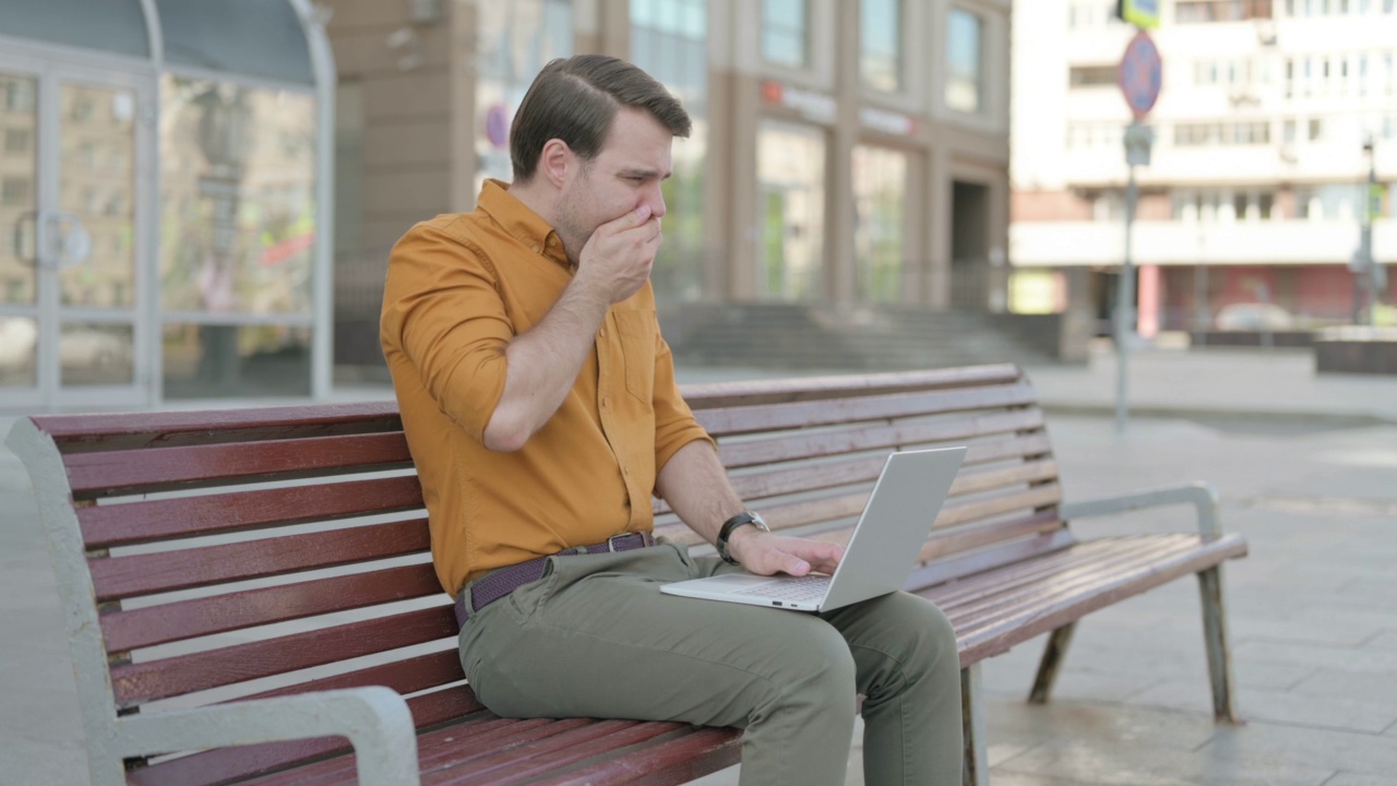 一名咳嗽的年轻人坐在户外长凳上使用笔记本电脑视频下载