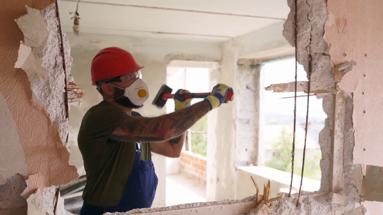 承包商用大锤破坏墙壁，打洞以便重新布置。一名男子正在用大锤子进行公寓改造的手工拆卸和拆除工作。穿着制服的建筑工人。视频下载