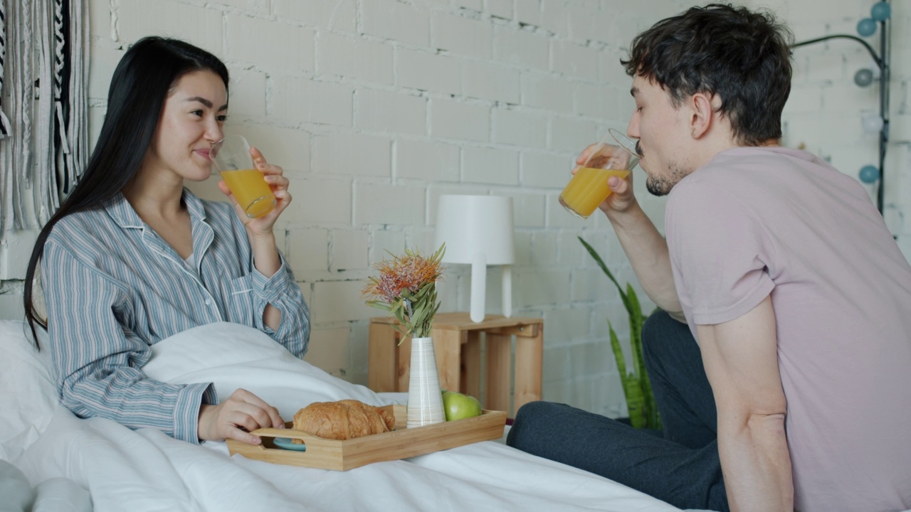 亚洲女士喂年轻的丈夫与羊角面包和喝果汁享受早餐在床上视频素材