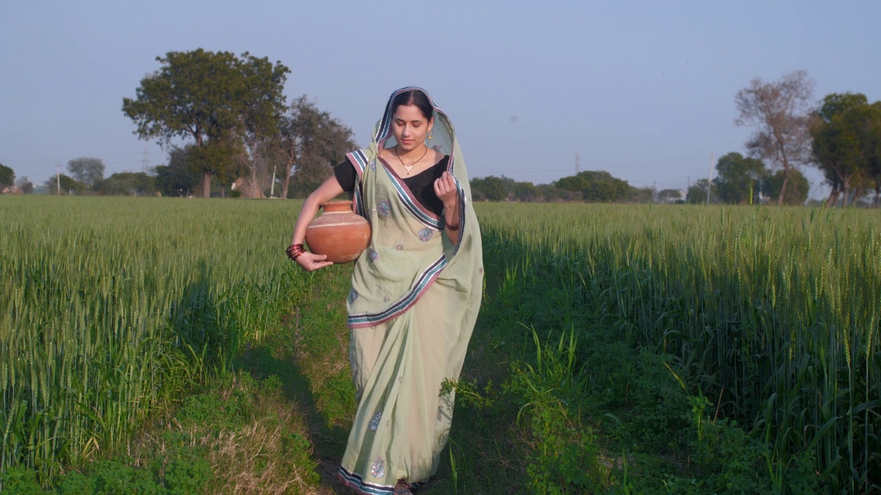 一个美丽的已婚家庭主妇拿着水罐走在田野上——村庄的景象视频素材