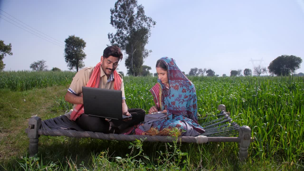 印度，一对年轻的乡村夫妇在他们绿色的麦田里——坐在帆布床上使用笔记本电脑，现代村民视频素材