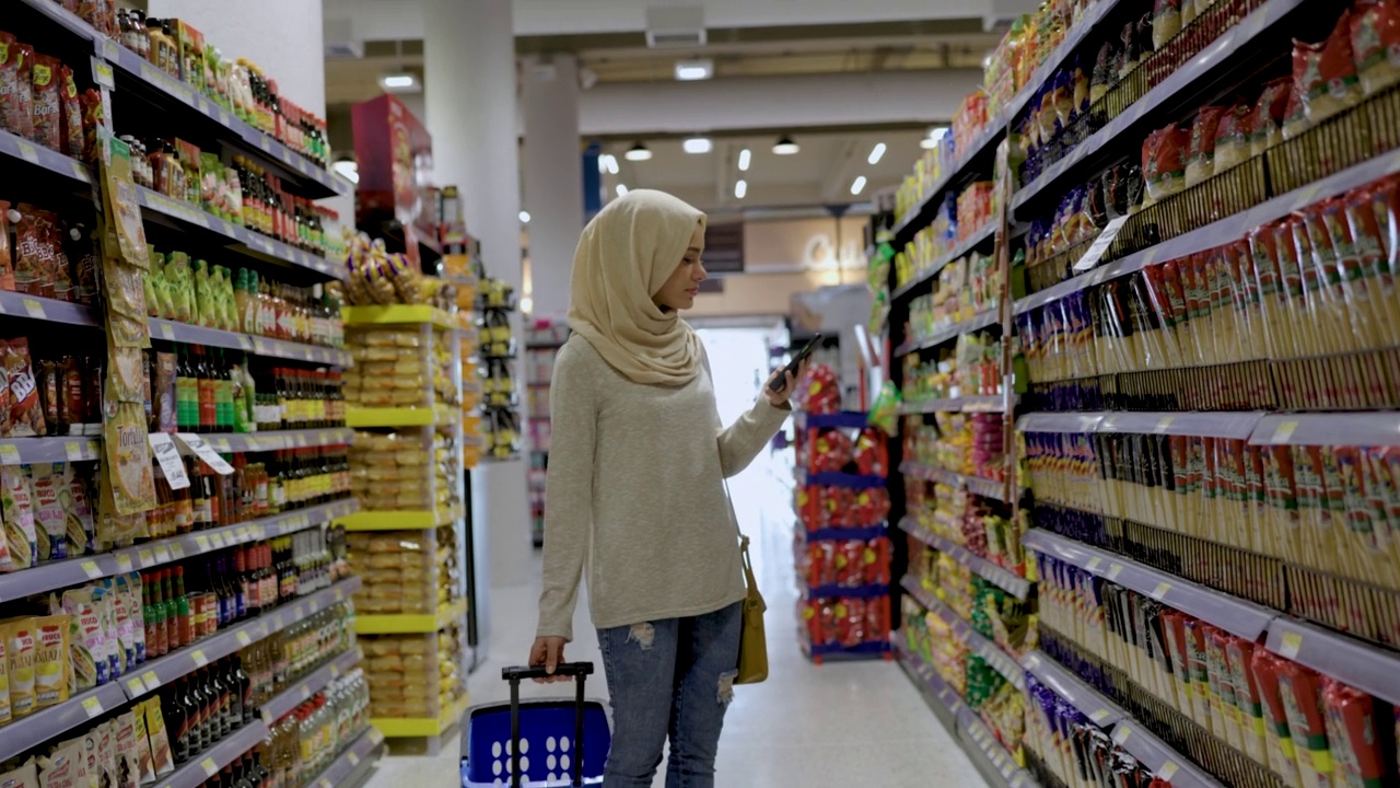 美丽的穆斯林妇女拉着一辆小推车，看着她的购物清单，同时走在超市的过道上，从货架上拿起一件产品视频素材