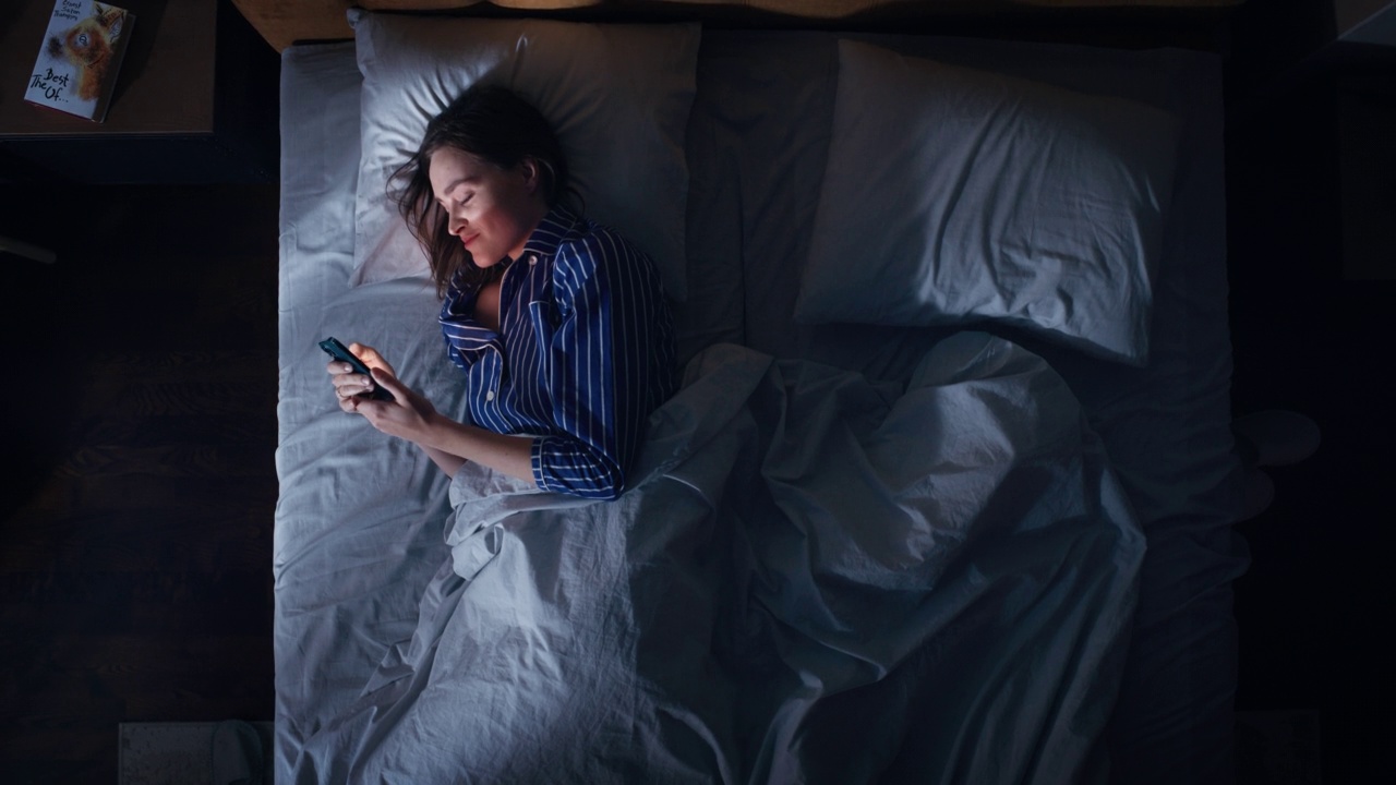 俯视图卧室公寓:美丽的年轻女子晚上在床上使用智能手机。微笑的女孩浏览社交媒体，浪漫的聊天，约会应用程序，做互联网网上购物的手机视频下载