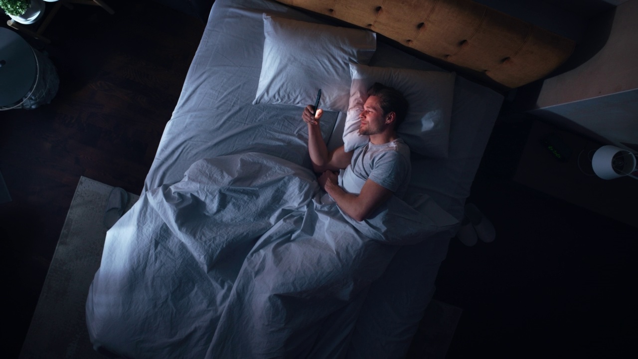 俯视图公寓卧室:英俊的年轻人晚上在床上使用智能手机。微笑的家伙浏览社交媒体，约会应用程序，做互联网在线购物的手机。上下缩放视频下载