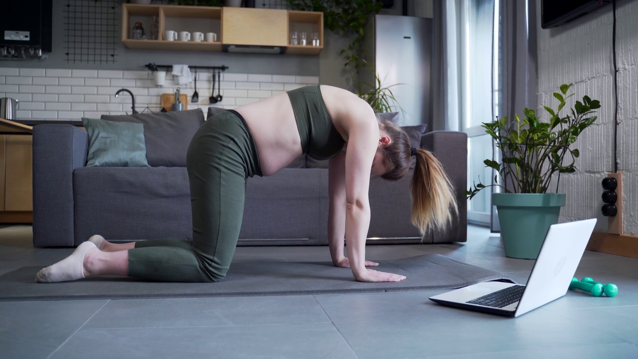 孕妇在家里的垫子上用笔记本电脑做体育锻炼。孕妇在线瑜伽课程。健身、体操、普拉提或在家庭视频课上拉伸。视频素材