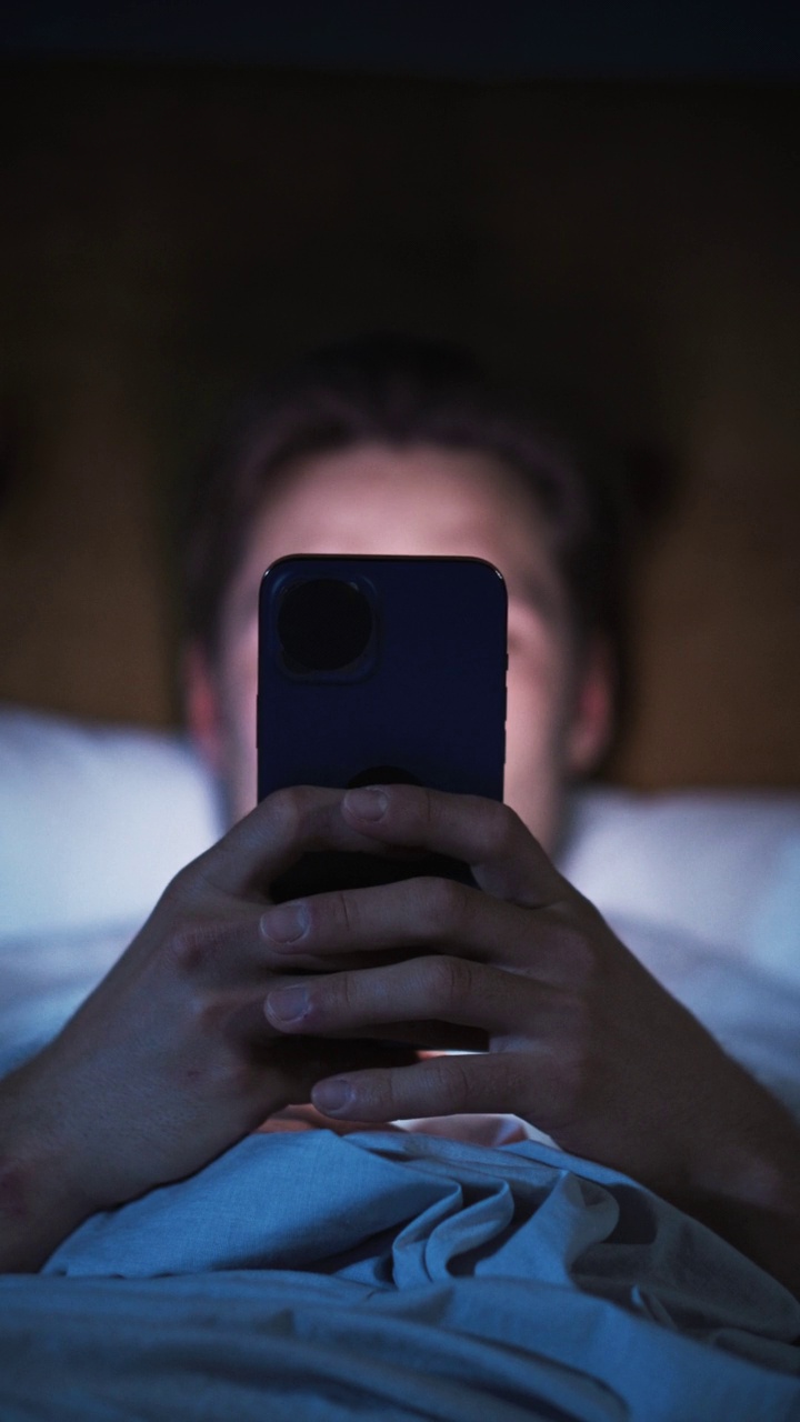 垂直屏幕:白人男性晚上在家床上使用智能手机。帅哥浏览社交媒体，阅读新闻，网上购物，深夜。拿着手机视频下载