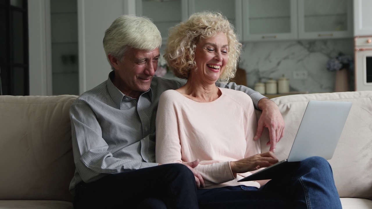白发苍苍的丈夫抱着坐在沙发上用笔记本电脑的妻子视频素材