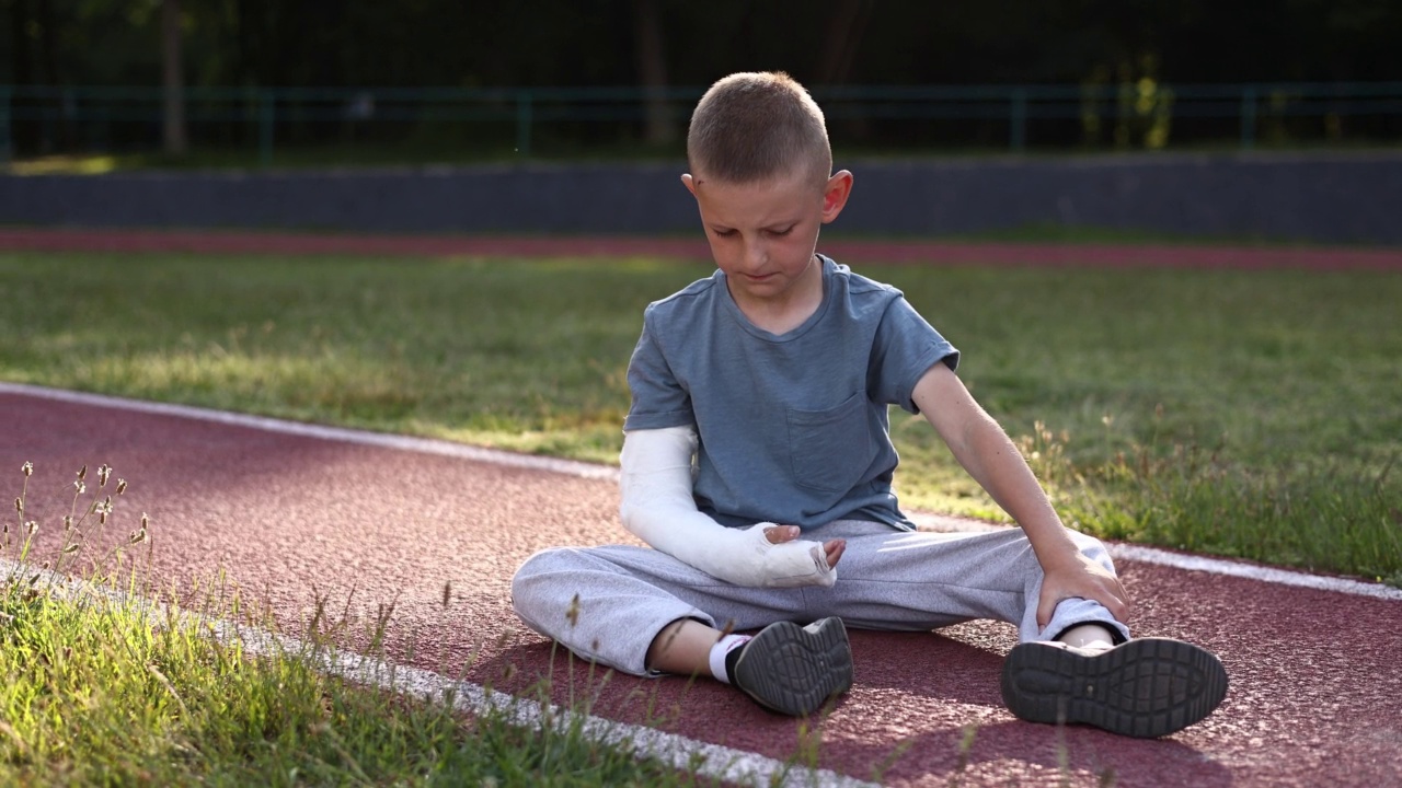 夏天，手受伤的小男孩坐在户外的操场上。9岁儿童骨折。骨折受伤。手臂上打了石膏视频下载