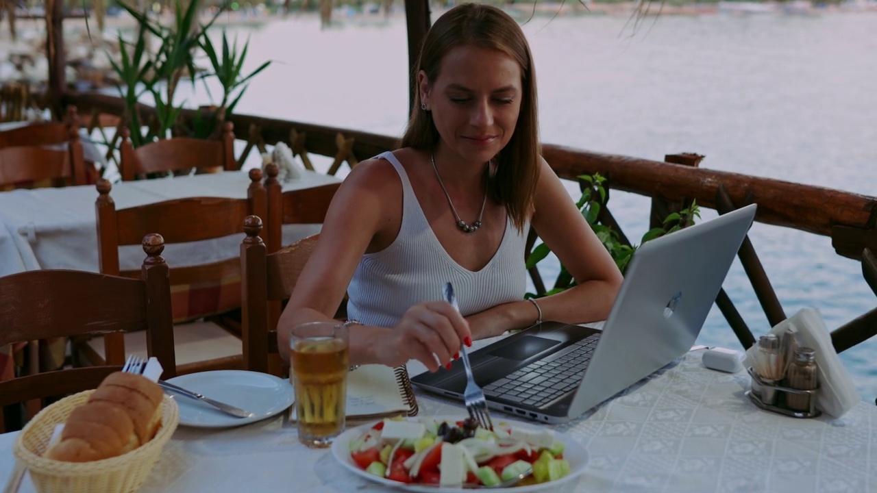 一名女子在海滨餐厅吃希腊沙拉，用笔记本电脑视频下载
