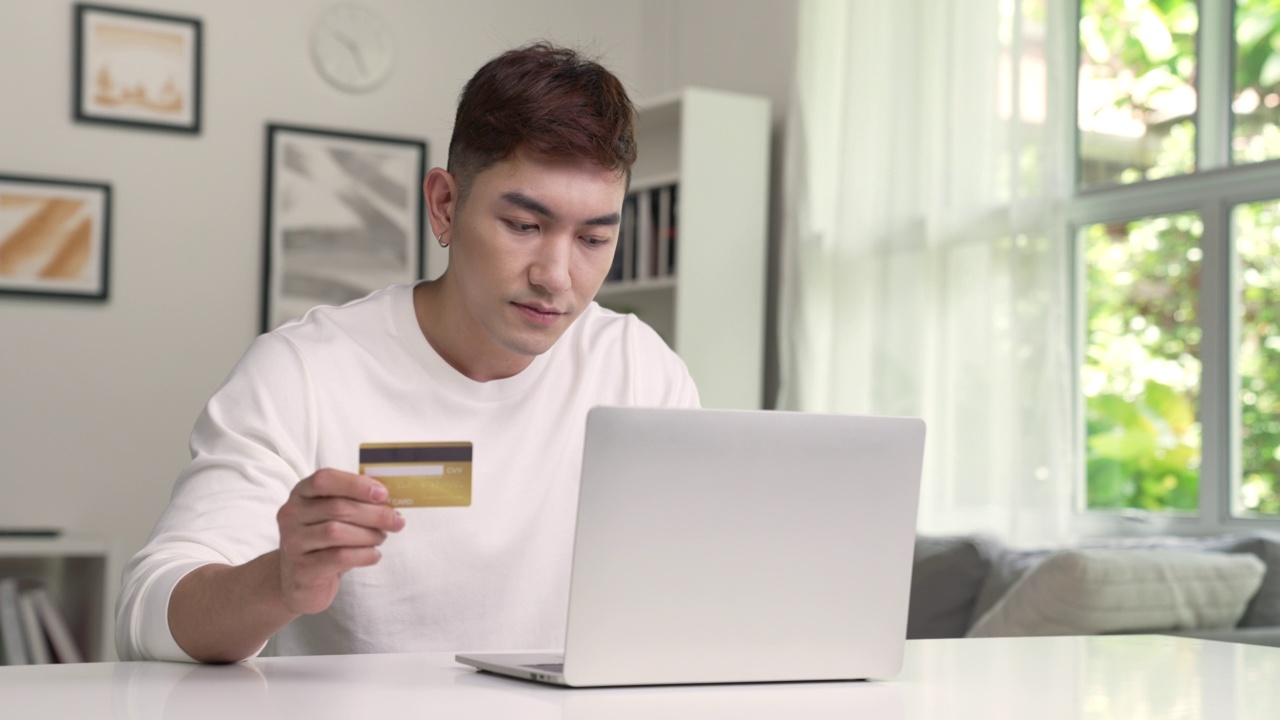 年轻的亚洲男子拿着信用卡，正在使用笔记本电脑。商人在家工作。网上购物，电子商务，网上银行，花钱，在家工作的概念视频素材