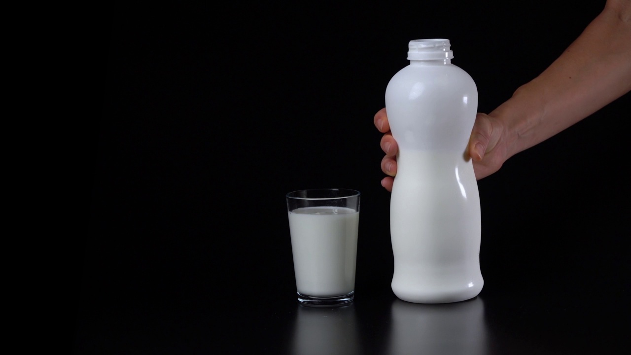 手从黑色背景的白色塑料瓶中倒入牛奶或开菲尔酒到一个玻璃杯子中。视频下载
