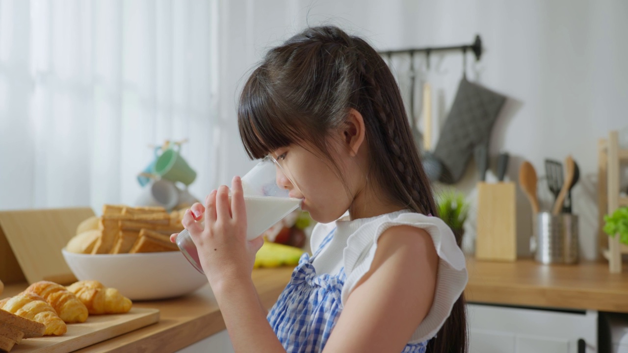 亚洲小孩拿着一杯牛奶在家里的厨房里喝。幼小的学龄前女孩或女儿喝牛奶，弄乱了牛奶的胡子和嘴唇后，在家里的医疗保健。视频素材