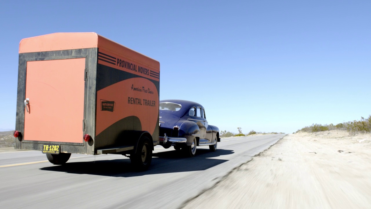 跟踪拍摄高角度下的1946年海军蓝色道奇自定义经典汽车在沙漠道路上行驶。汽车拖着橙色的租赁拖车。镜头摇摄到汽车的乘客侧视频下载