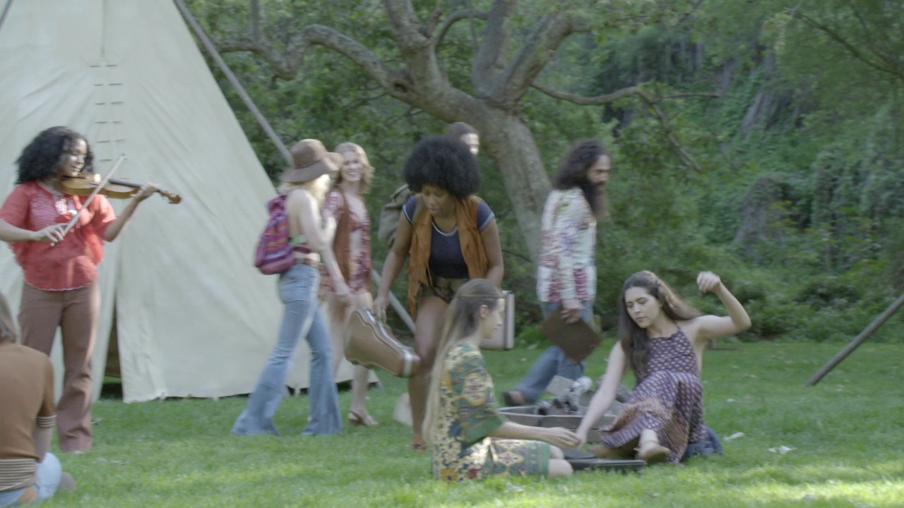 从右到左中间角度拍下人们背着乐器箱走出帐篷或圆锥形帐篷的画面。女人坐在田野的草地上。可能是夏令营。人们穿着那个时代的衣服。可能是嬉皮士视频下载