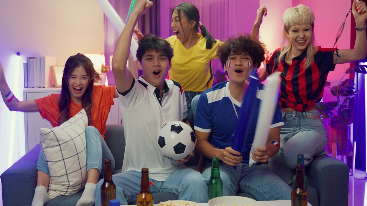 一群快乐的亚洲青少年坐在沙发上看欢呼体育，足球比赛，世界杯在电视上在晚上。视频素材