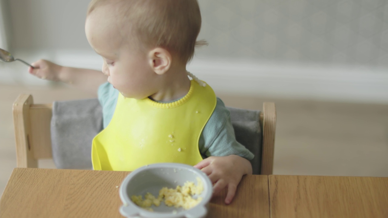 穿着黄色围兜的婴儿抱着勺子坐在餐桌旁学习进食视频素材