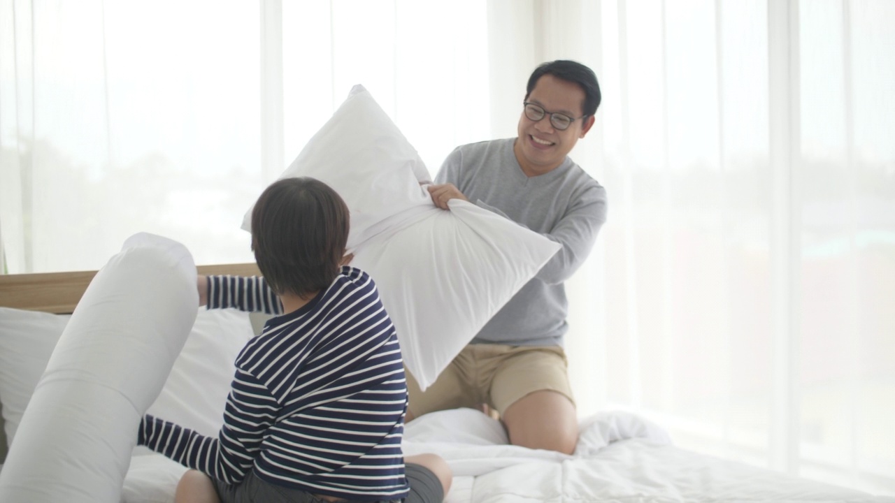 快乐的家庭时间，年轻的亚洲父亲和儿子一起在床上玩打枕头在卧室快乐视频素材