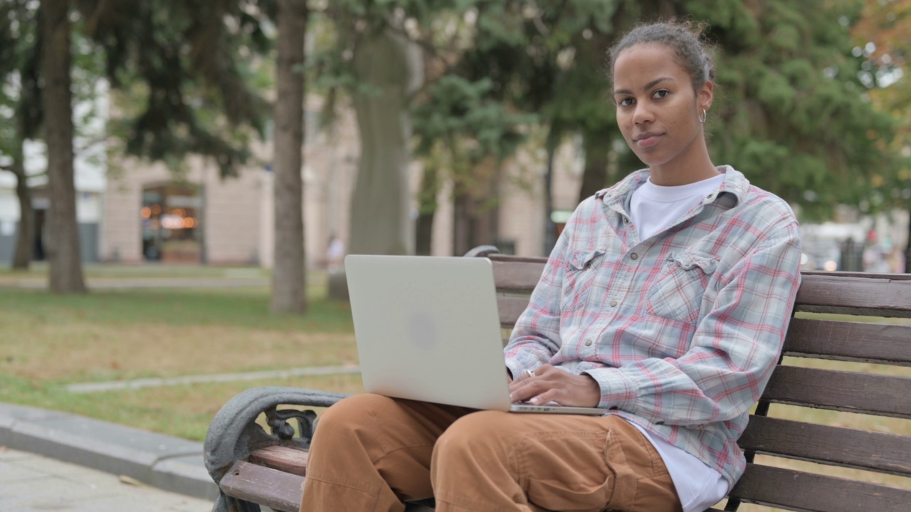 非洲妇女坐在户外的长凳上，用笔记本电脑看相机视频素材