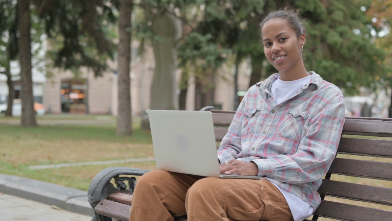 带笔记本电脑的非洲妇女坐在户外的长凳上对着相机微笑视频素材