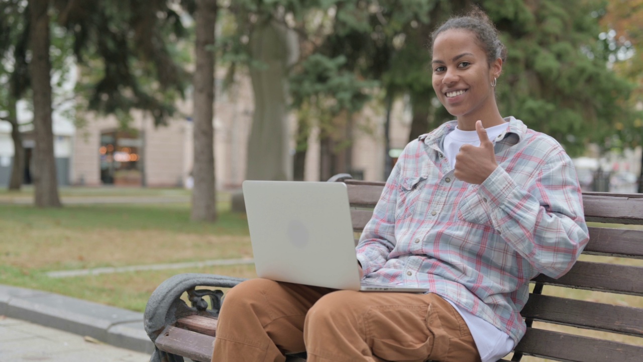 一名非洲妇女坐在户外的长凳上，用笔记本电脑翘起大拇指视频素材