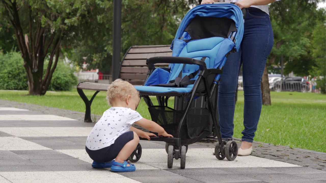 夏天，妈妈带着孩子在公园散步，阳光明媚的日子里，孩子带着婴儿车在人行道上玩耍。视频下载
