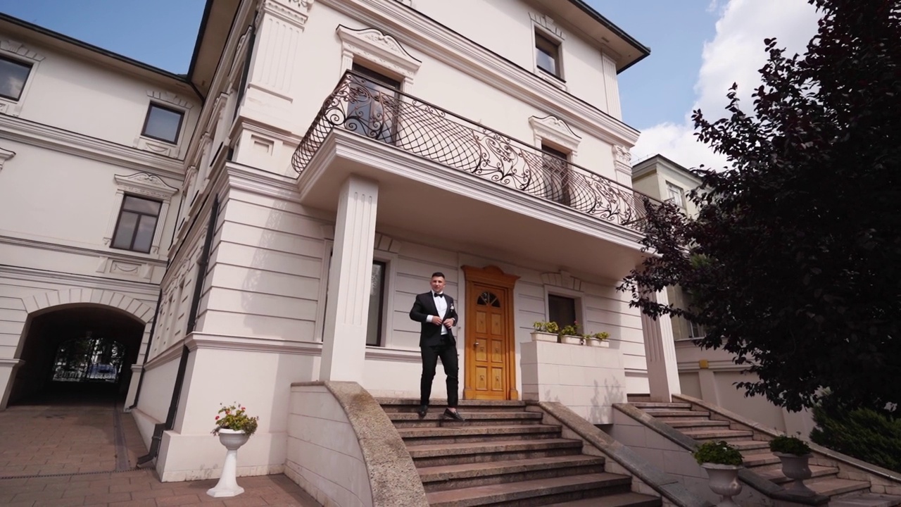 一个穿着黑色西装、打着领结的专横男人走下大理石楼梯，走出他的豪华豪宅。百万富翁正走出他的宫殿。新郎以慢镜头到达婚礼现场。视频素材
