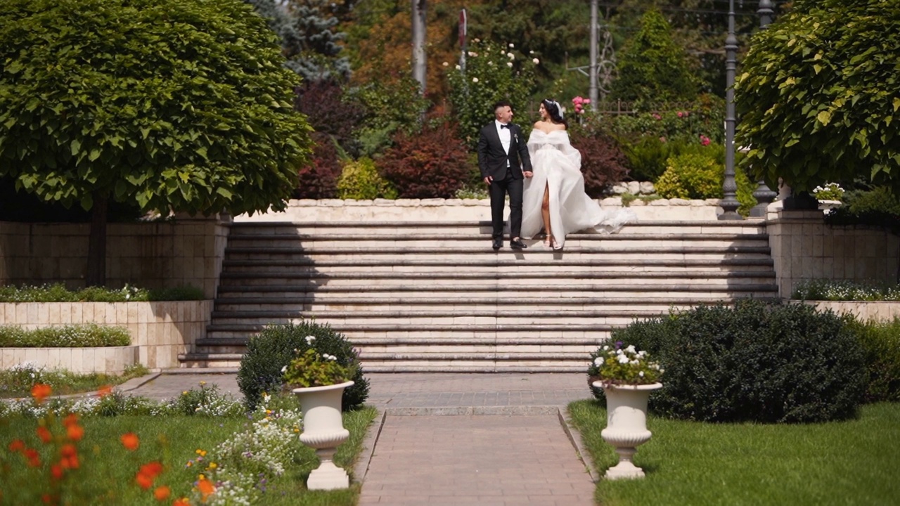 新娘和新郎走下豪华豪宅院子里的大理石楼梯。白衣长纱的男女手牵着手在宫廷花园漫步。新婚夫妇去婚礼场地参加仪式。视频素材