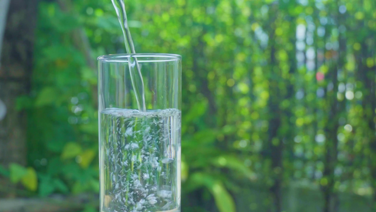 在一片模糊的绿叶背景下，将纯净水倒入一杯水中视频素材