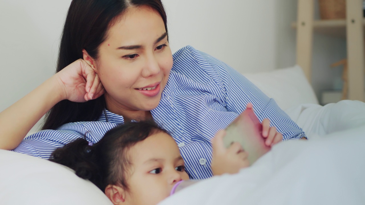 亚洲妈妈和宝宝躺在床上看智能手机视频素材