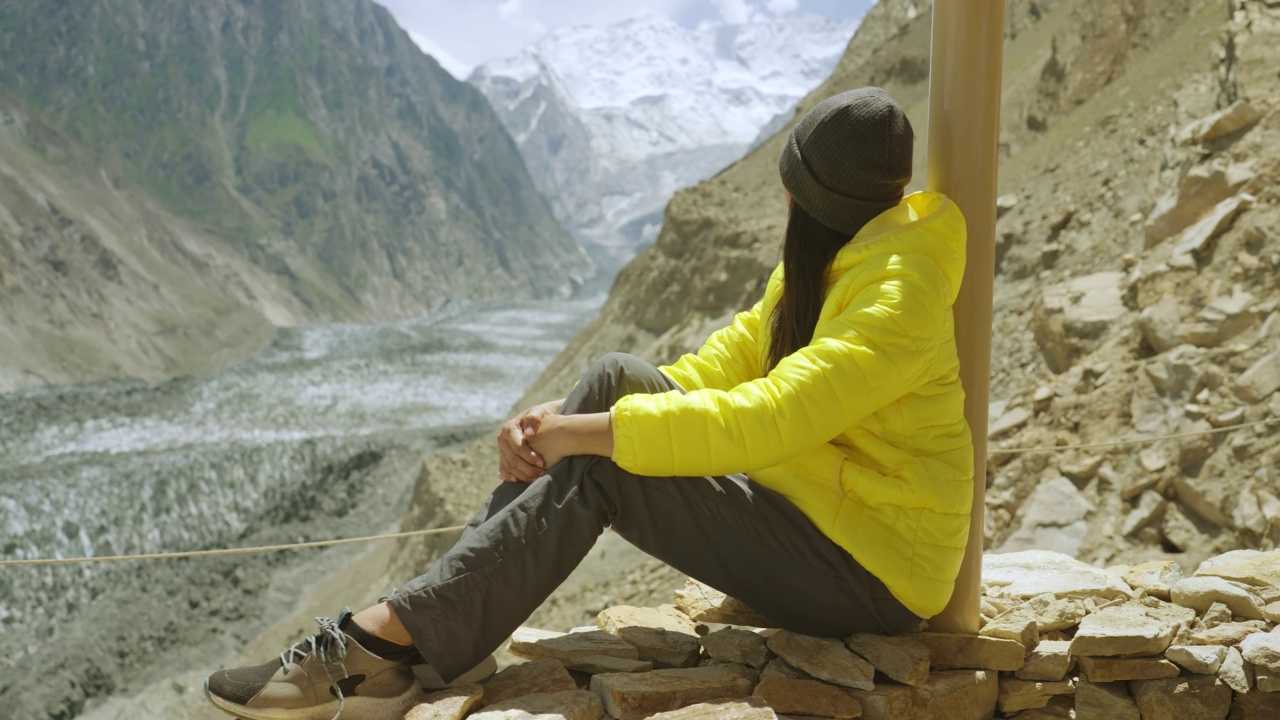 自由旅行者年轻女性穿着冬季夹克黄色坐着，看着冰川的景色在气候变化。种族多样化的旅行者概念。与α香奈儿。视频素材