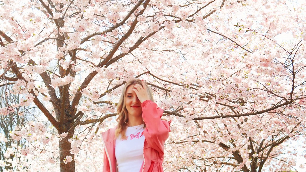 年轻女子微笑在盛开的樱花或樱桃树在春天穿粉红色和白色的衣服视频下载