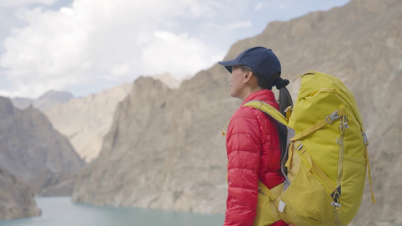 后视图，自由的旅行者的快乐年轻的女人穿着冬季夹克红色，背着黄色的背包站在湖边享受旅行假期的晴朗的天空在早上。种族多样化的旅行者概念。与α香奈儿。视频素材