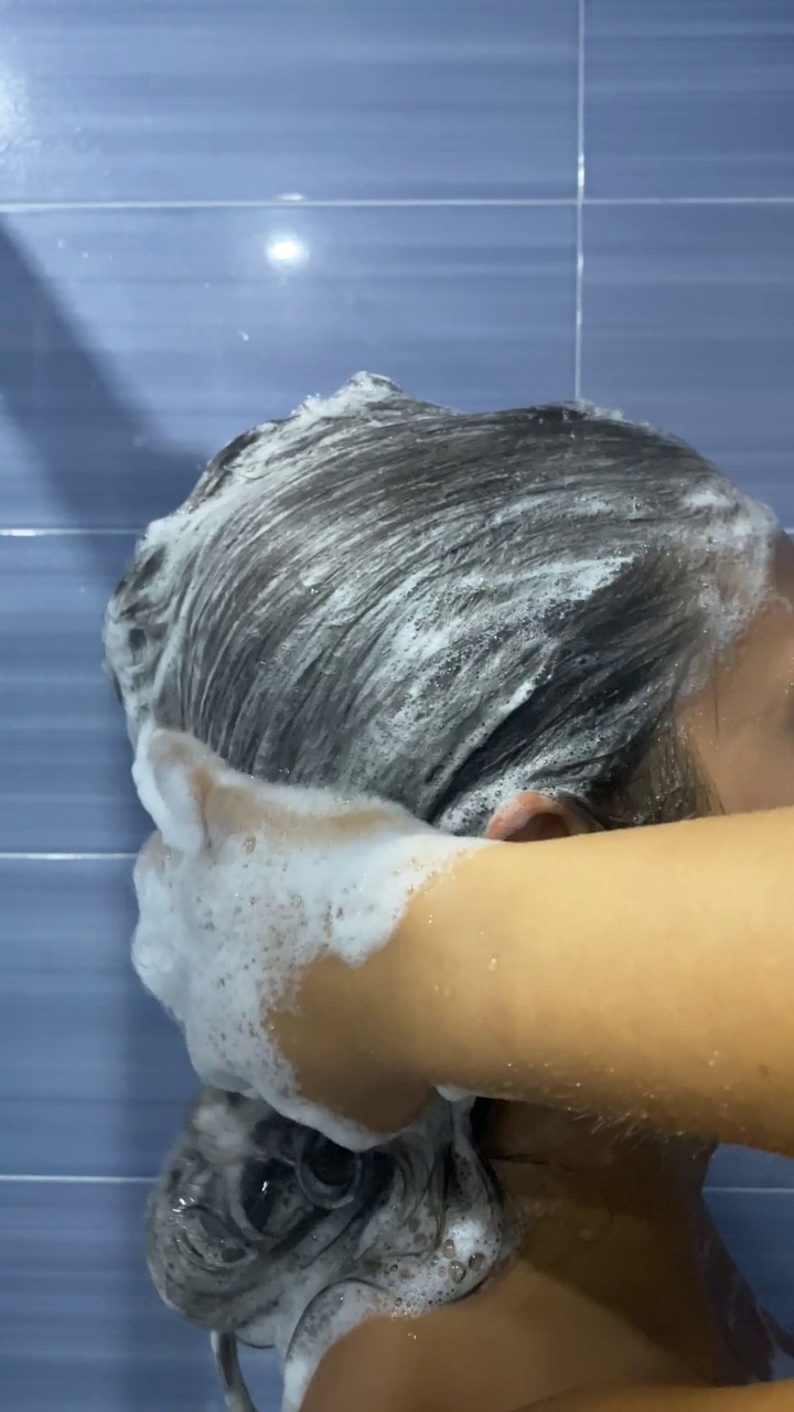 淋浴中的女人在用泡沫洗发露将头发涂满泡沫后冲洗头发。一个小女孩在淋浴时用慢动作从头发上取下肥皂的垂直视频。视频素材