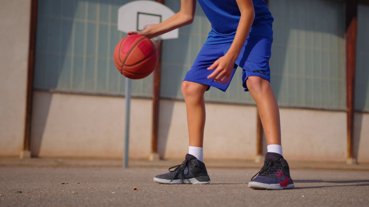 活跃的男性篮球运动员在小区街球训练期间，在清晨升起的太阳的光线下，在室外球场练习持球技巧，盘带球之间的腿视频下载