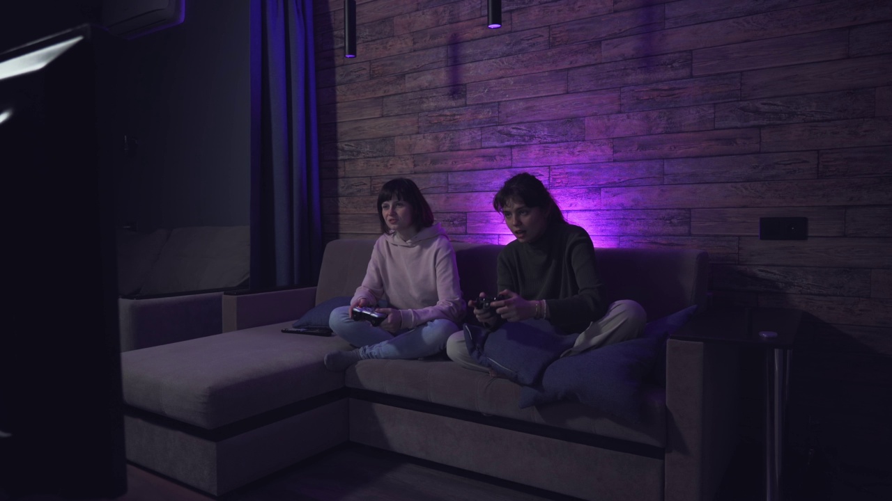两个年轻女子坐在沙发上玩游戏机视频游戏手柄手柄。女性游戏玩家在家里一起开心地笑着在线娱乐视频素材