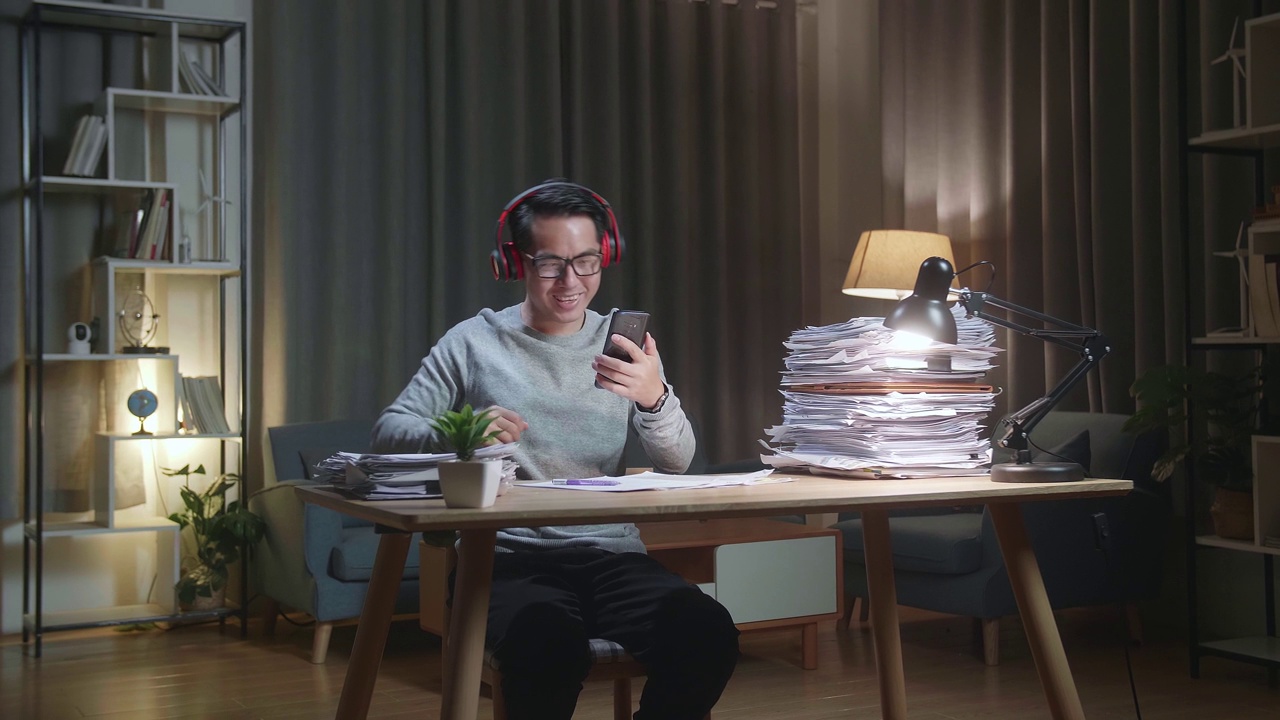 亚洲男子在家处理文件后，戴着耳机在智能手机上听音乐视频下载
