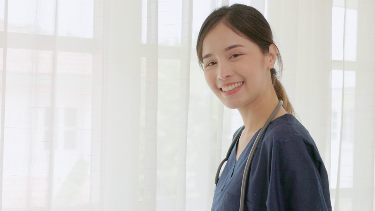 看着镜头的是一个快乐的年轻亚洲女医生，穿着白色的医疗外套，拿着听诊器。微笑的女医生在医院办公室。美丽的医院住院医生微笑着视频素材