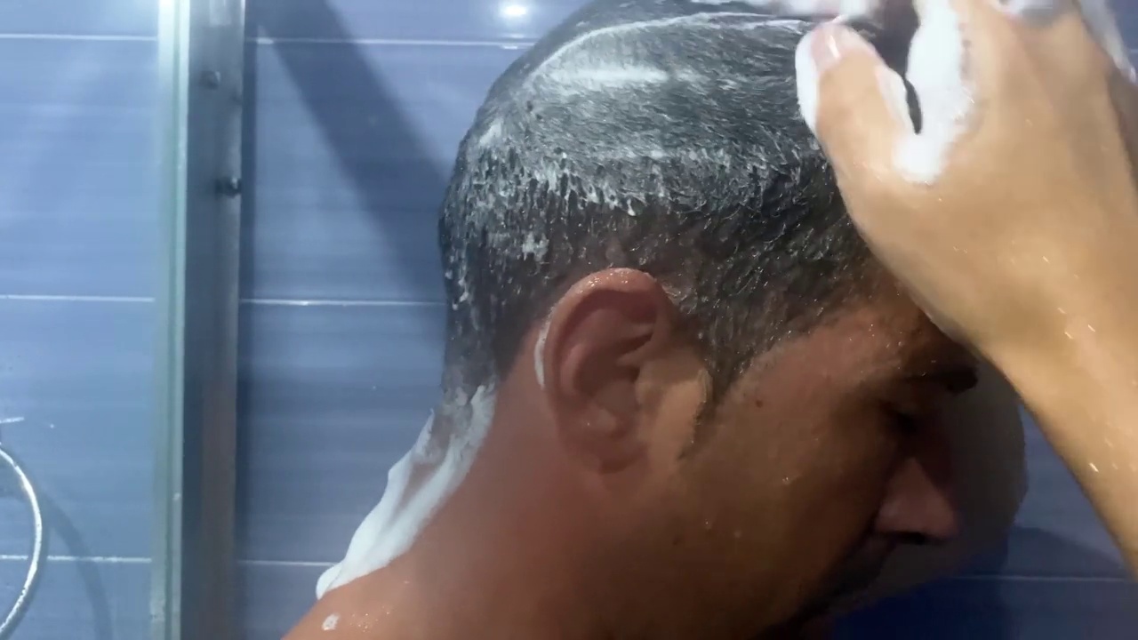 一个男人在浴室里揉着他的头发，用泡沫洗发露在上面起泡沫。一名年轻男子在淋浴时用泡沫抹头发的视频。视频素材
