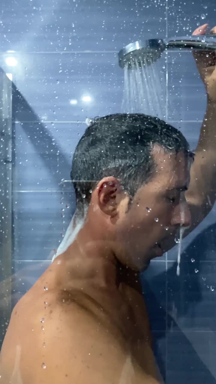 一个男人在浴室里用泡沫洗发露冲洗头发。一名年轻男子在淋浴时用慢动作从头发上去除肥皂的垂直视频。视频素材