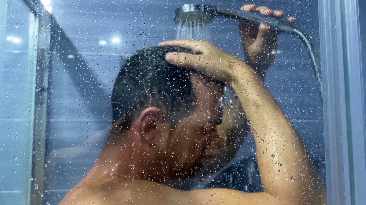 一个男人在浴室里用泡沫洗发露冲洗头发。一名年轻男子在淋浴时用慢动作去除头发上的肥皂的视频。视频素材