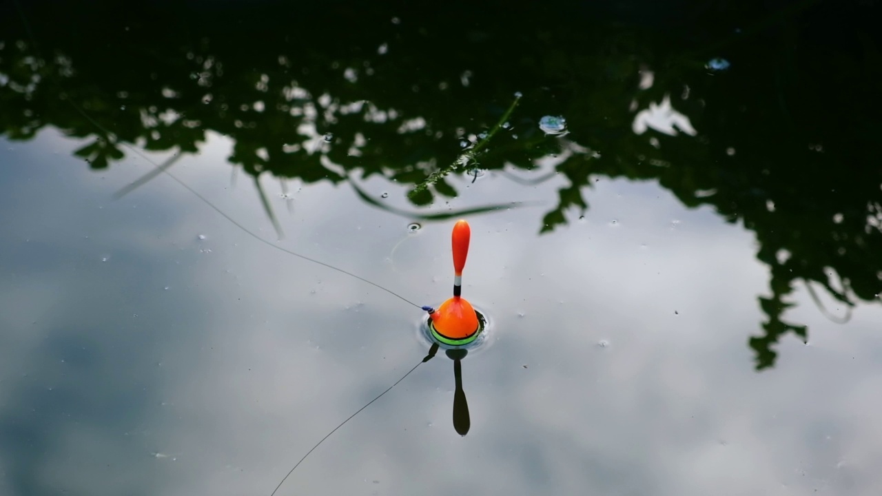 浮，钓线在湖面上。钓鱼。明亮的橙色钓鱼浮在黑暗的池塘咬。一只浮标在蓝色的水面上转着圈。前视图。爱好和放松。视频下载