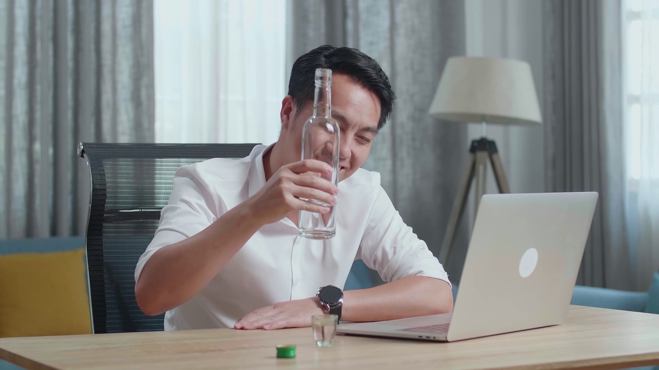 一名亚洲男子在家里用笔记本电脑进行视频通话时一边欢呼一边喝伏特加视频下载