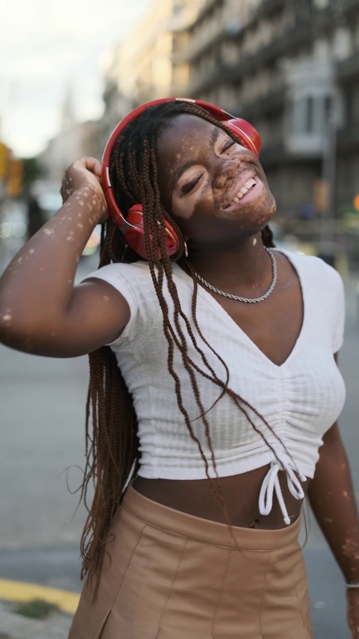 患白癜风的非裔美国妇女戴着耳机在街上听音乐时微笑。视频下载