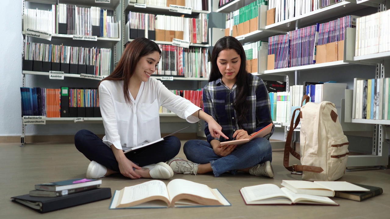 两个拉丁女学生坐在图书馆的地板上，手里拿着书视频素材