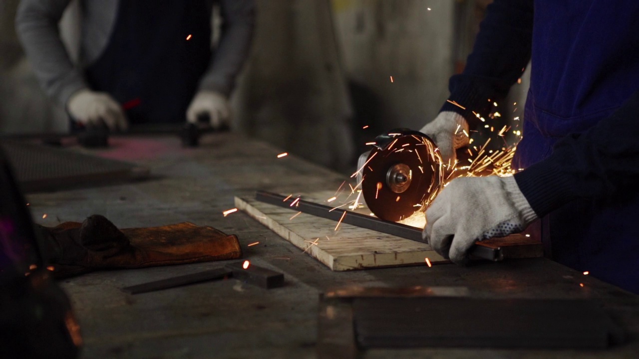 戴着防护手套的金属工人用角磨机在工作台前慢动作切割一块金属，他的同事在背景中工作的手的近景侧面视频下载