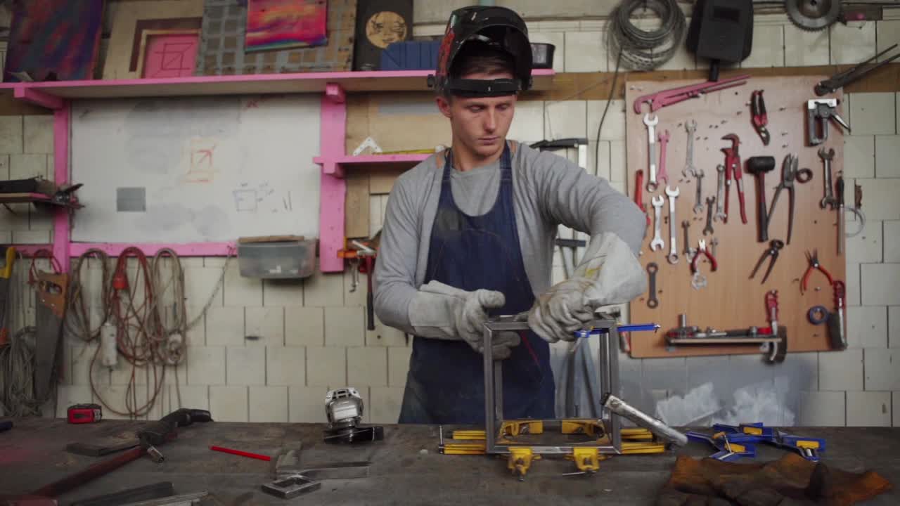 慢动作中镜头年轻的金属工人戴着安全面具和手套使用角磨机磨钢立方体和焊枪连接它站在车间的工作台边视频下载