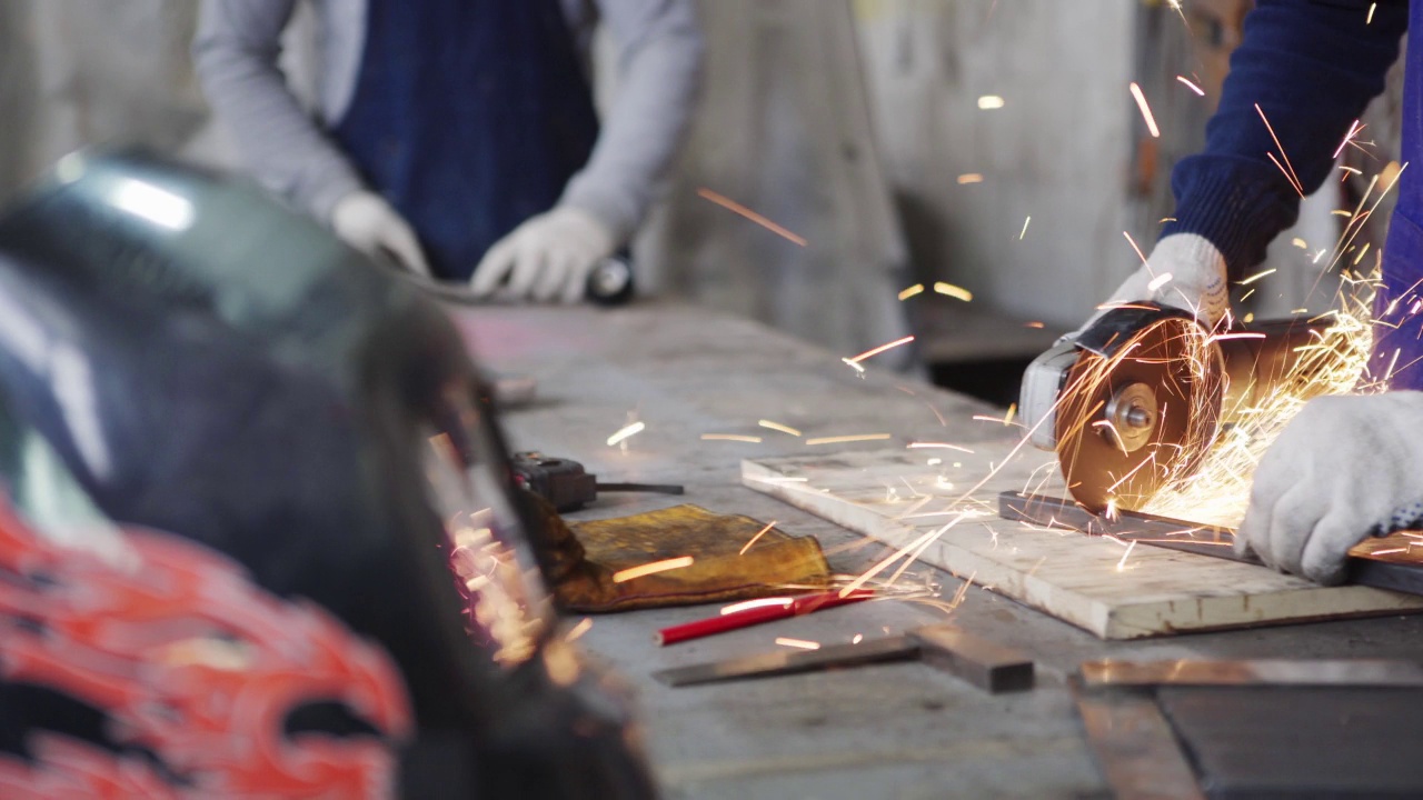 特写跟踪镜头的手，熟练的金属工人在保护手套切割一块金属与角度磨床站在工作台，他的同事在背景工作视频下载