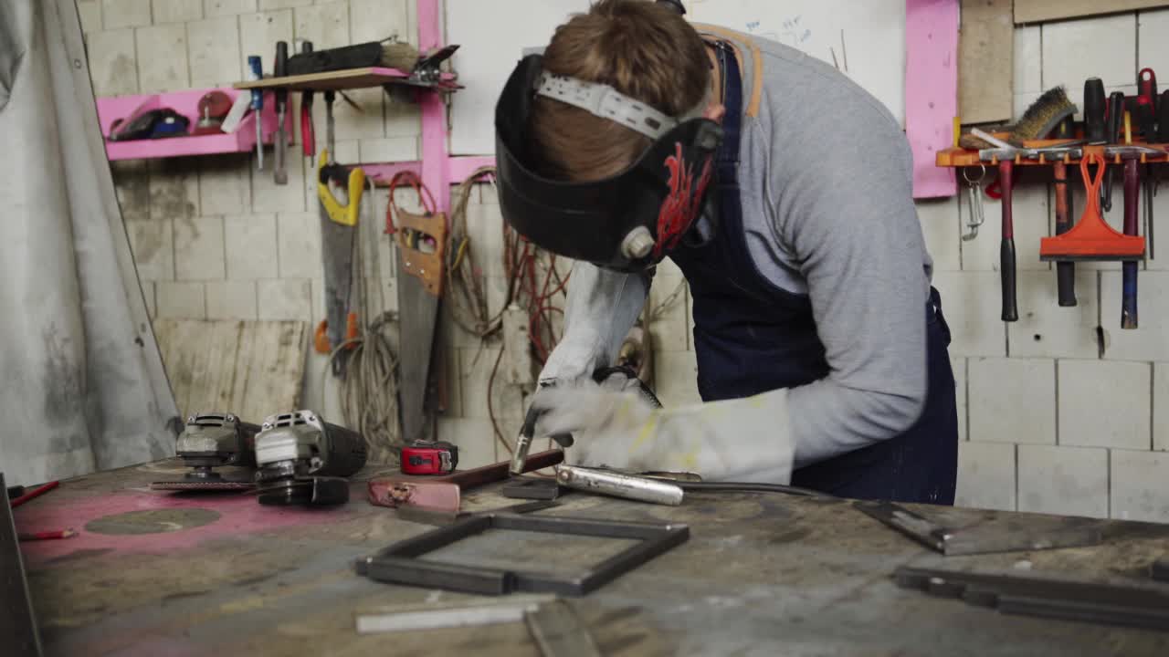 中景:年轻的金属工人戴着安全面具和手套，用焊枪站在车间的工作台前焊接金属视频下载